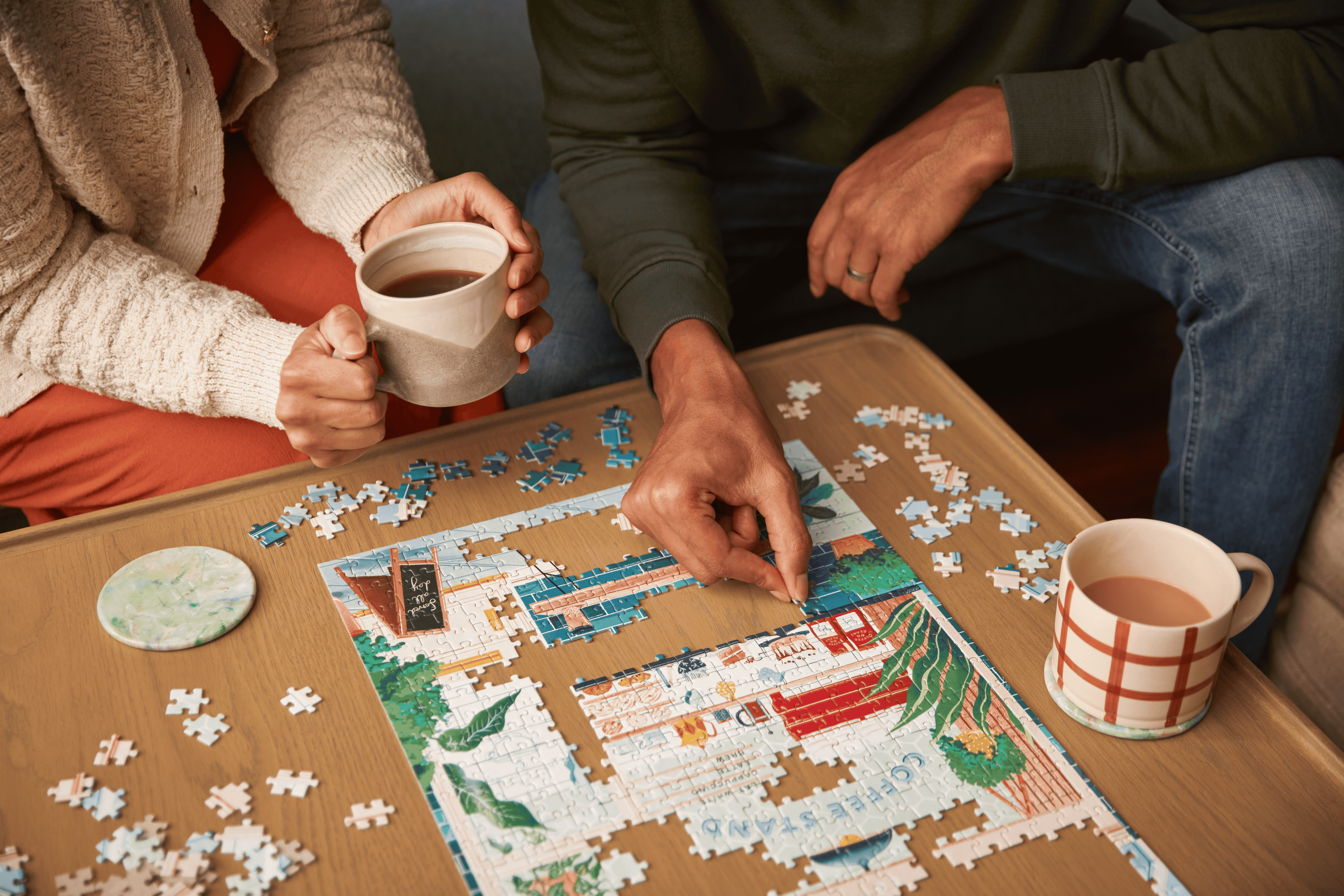 Overhead shot of couple doing a 500-piece jigsaw together with warm drinks in hand.