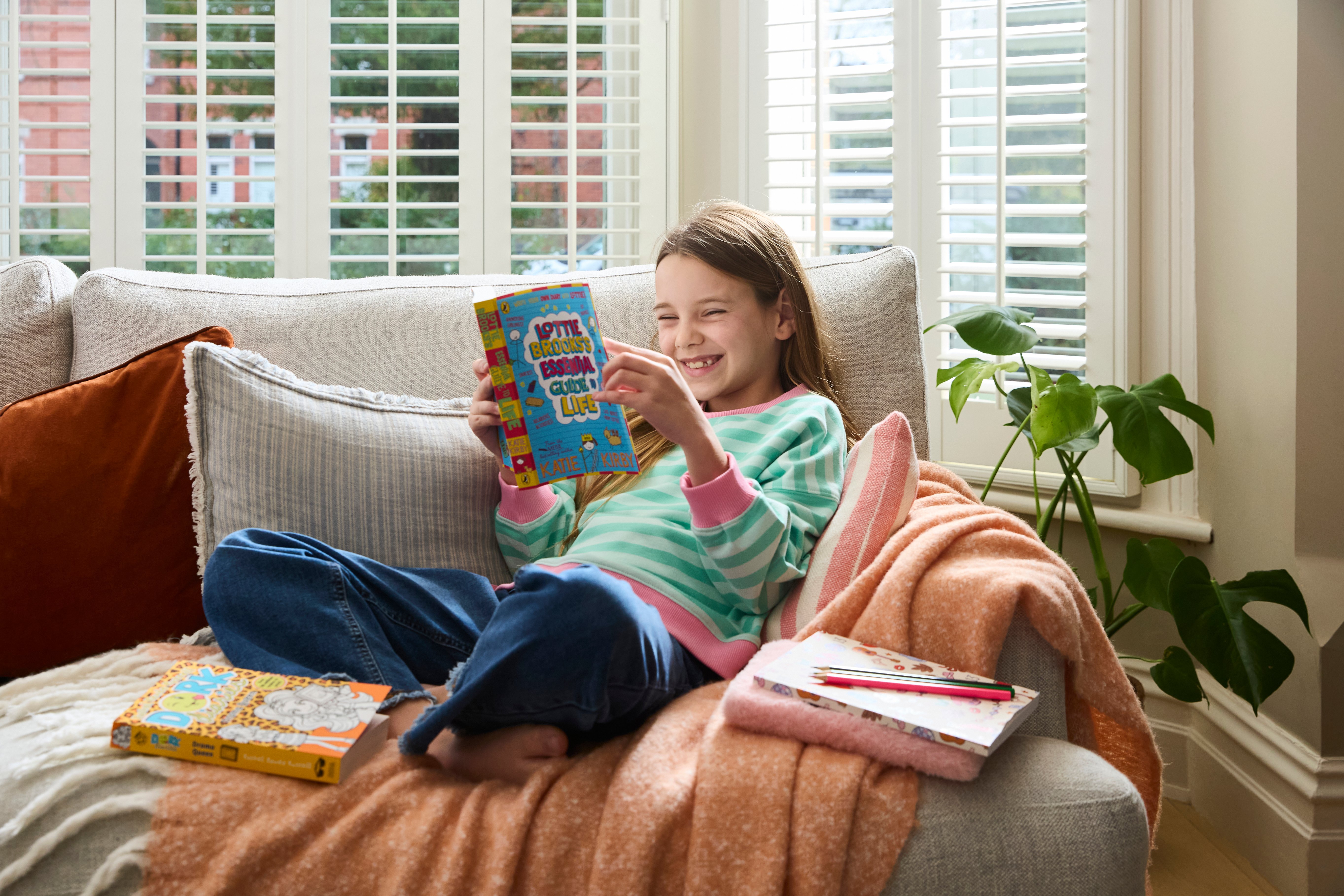 Find Their Story: The Best Kids’ Books by Interest A girl sits on a sofa with a bright smile as she reads Lottie Brooks.