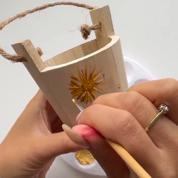 A fine paint brush is painting a large flower on a wooden bucket.