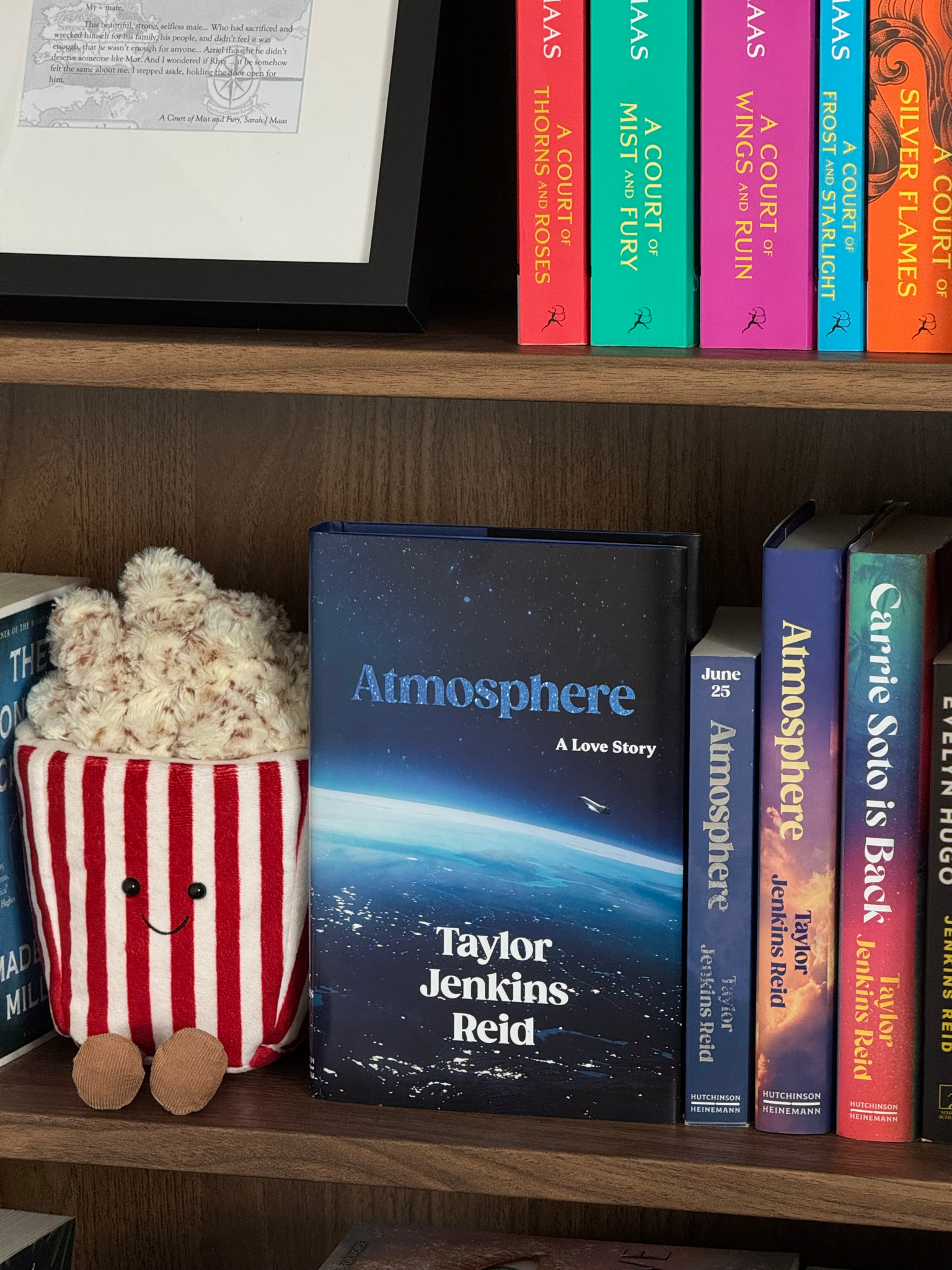 Atmosphere sits on a dark wood book shelf next to a popcorn jellycat and other books by Taylor Jenkins Reid.