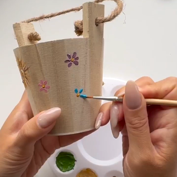 A fine paint brush is painting small flowers on a wooden bucket.