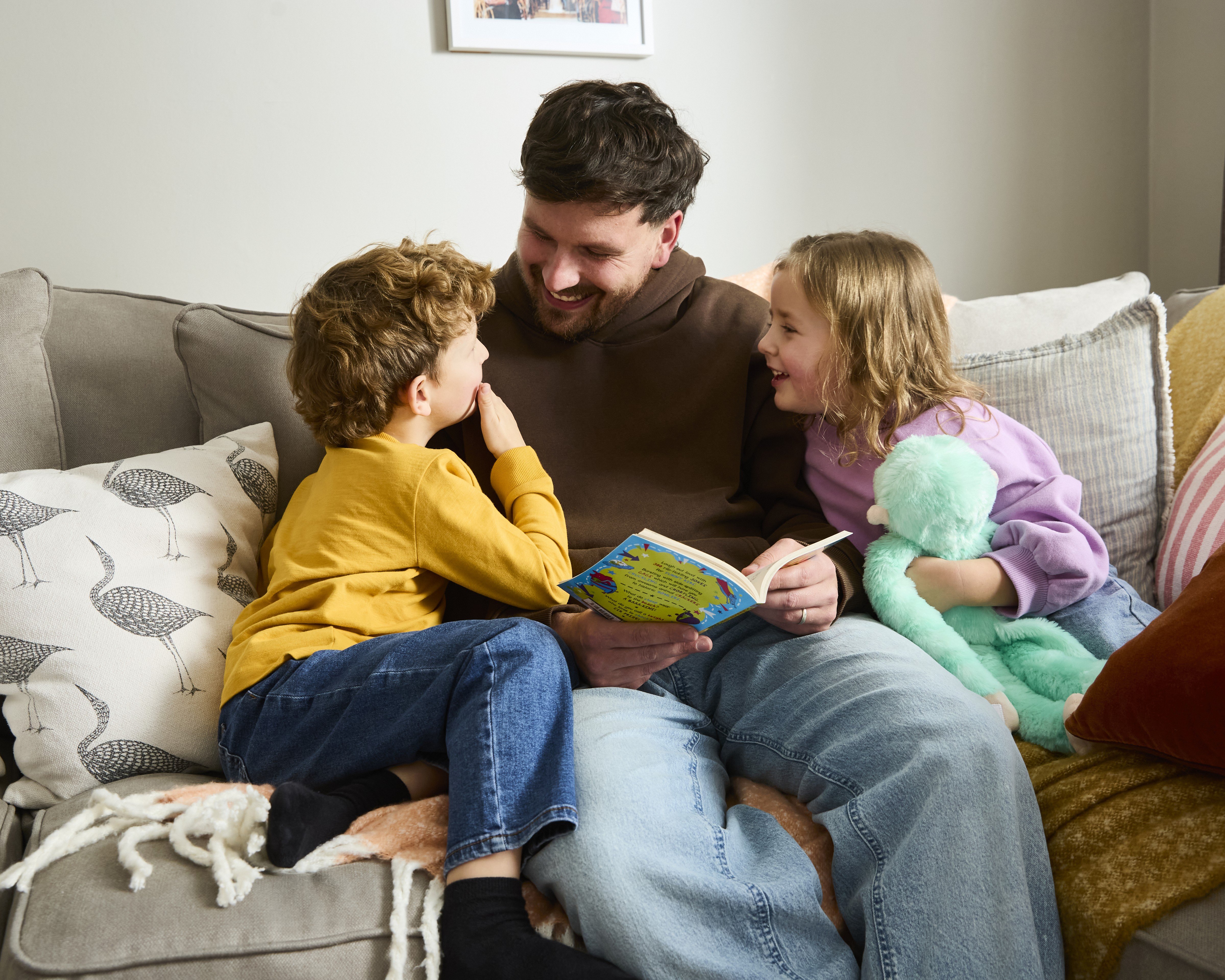 Two children read with Dad on the sofa. All are smiling at each other.