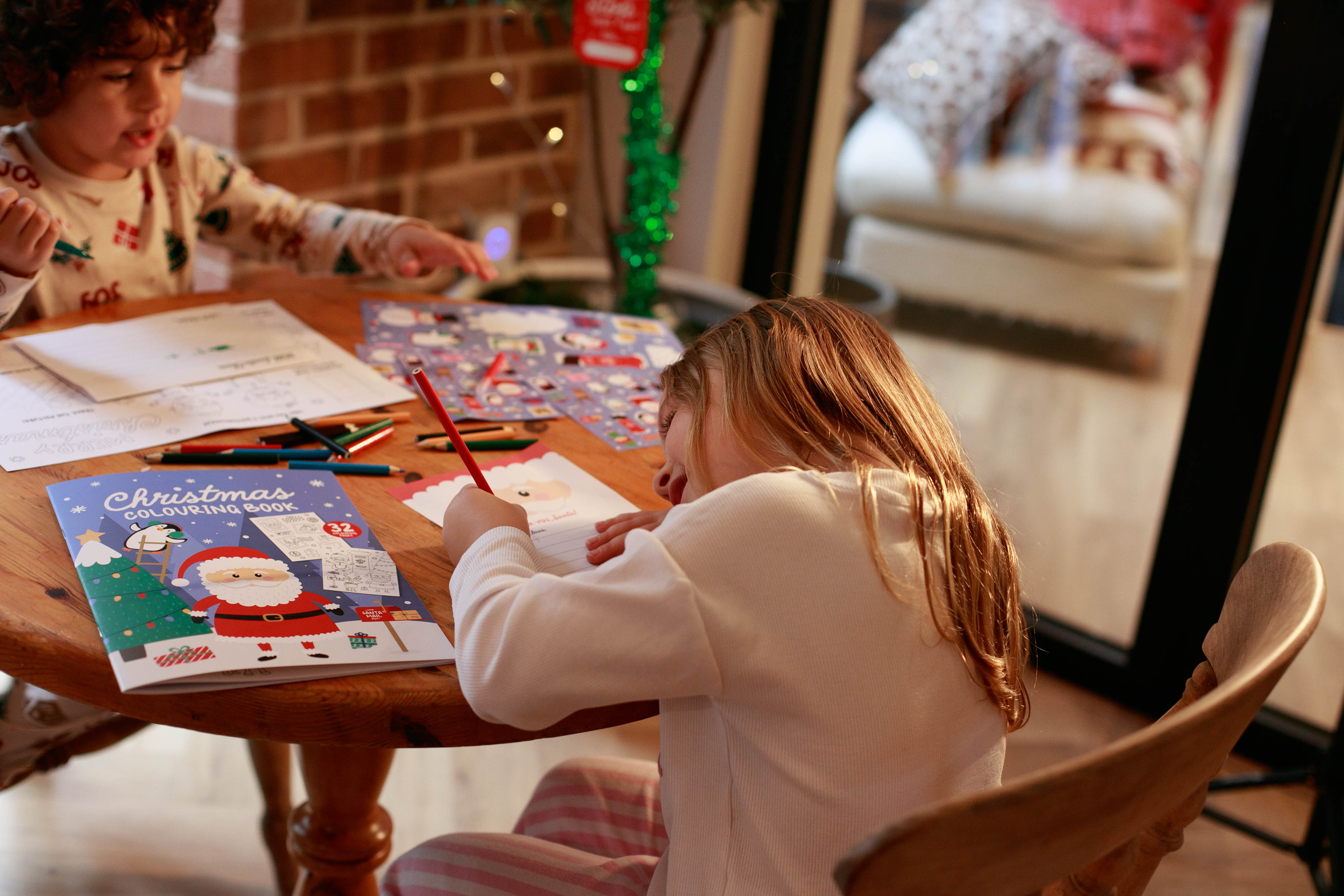 A young girl is sat at the table with her brother colouring and writing a letter to Santa.