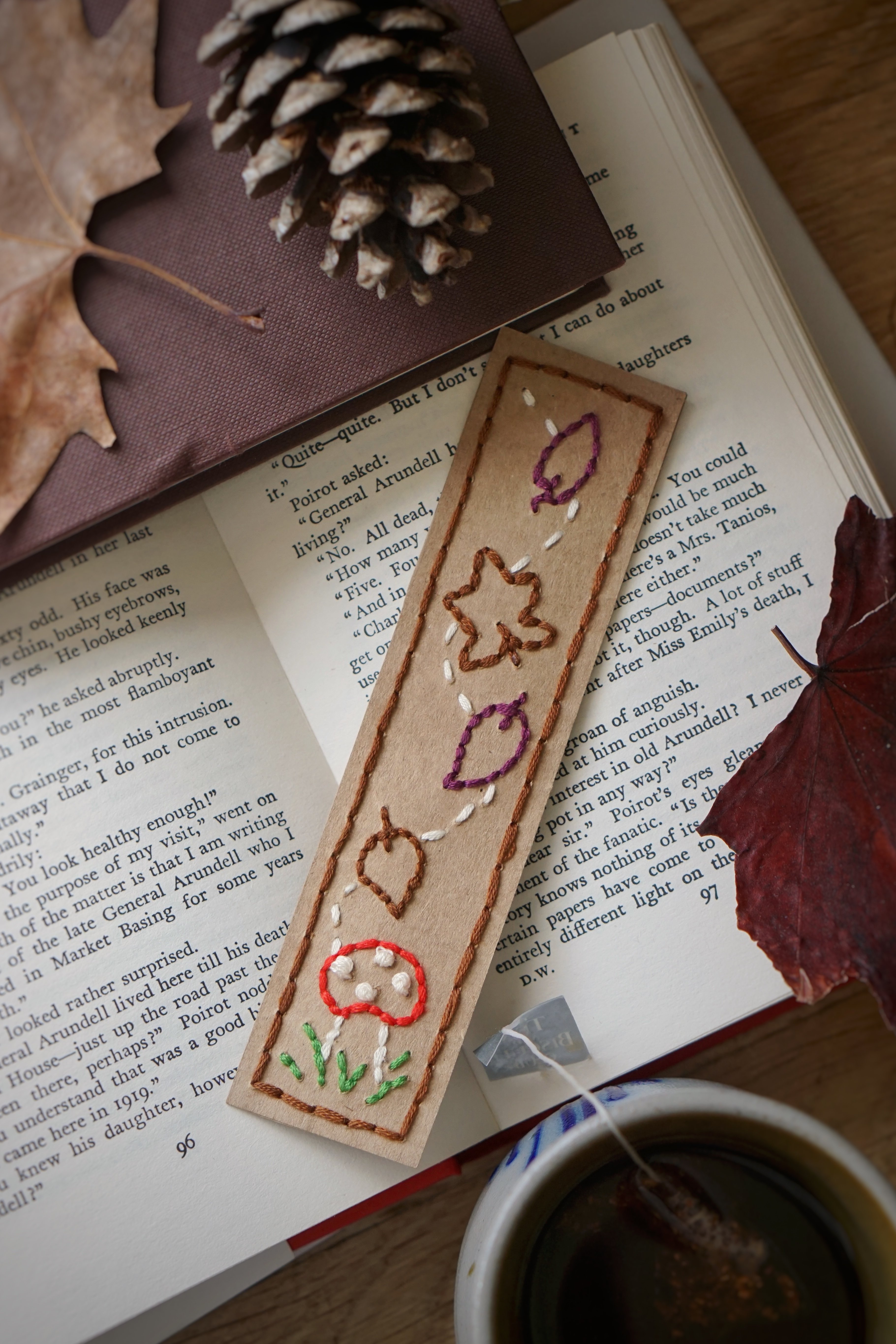 DIY autumn embroidered bookmark sits on an open book with some autumn decor around it and a hot drink.